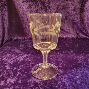 May include: A clear glass wine glass with a delicate etched design of white leaves and vines. The glass has a tall, slender stem and a round base.
