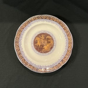 May include: A white ceramic plate with a gold and purple border. The center of the plate features a brown and gold image of a knight on horseback fighting a dragon.