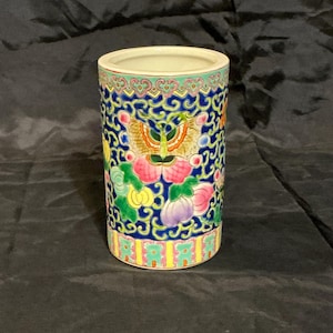 May include: A cylindrical ceramic vase with a detailed floral and bird design. The vase features a blue background with colorful flowers, birds, and intricate patterns. The top and bottom rims have decorative borders with a mix of colors. The vase is approximately 5 inches tall.