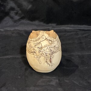 May include: A small, rounded ceramic vessel with a cream-colored base and a lid. The upper portion features intricate, raised brown and tan relief carvings of tangled, root-like or organic textures that wrap around the top of the container.