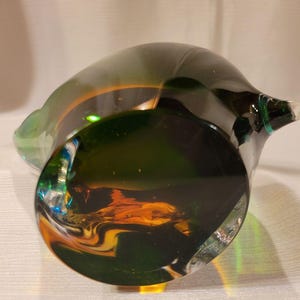 May include: A hand-blown glass sculpture with a dark green base and a lighter green rim. The interior features swirls of orange and yellow. The piece has a unique, organic shape and a clear, smooth finish.