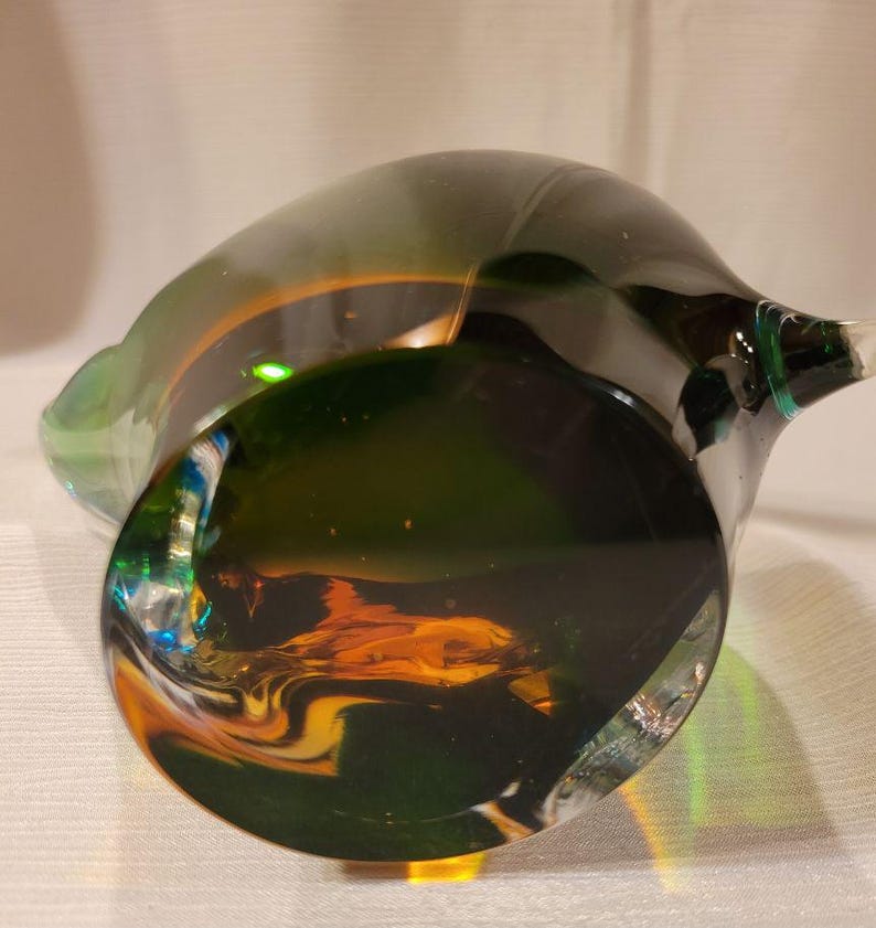 May include: A hand-blown glass sculpture with a dark green base and a lighter green rim. The interior features swirls of orange and yellow. The piece has a unique, organic shape and a clear, smooth finish.