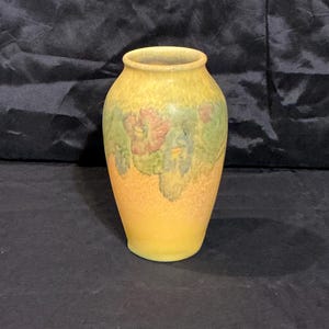 May include: A ceramic vase with a rounded body and a narrow neck. The surface features a gradient of yellow and orange tones, accented by a decorative band of muted green and reddish-brown floral patterns encircling the upper portion of the vessel.