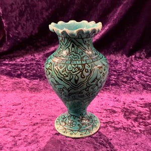 May include: A turquoise ceramic vase with a scalloped rim and intricate black floral designs. The vase has a flared base and stands on a purple velvet surface.