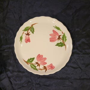 May include: A white ceramic plate with a scalloped edge, decorated with pink floral designs and green leaves. The plate has a simple, elegant design, perfect for serving food. The plate is approximately 10 inches in diameter.