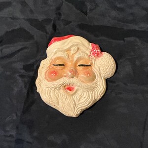 May include: A vintage Santa Claus face ornament. The ornament features a white beard, rosy cheeks, and a red hat. The eyes are closed, and a small pink flower is on the side of the hat. The ornament is made of a textured material.