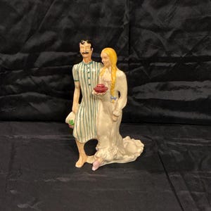 May include: A ceramic figurine depicting a couple. The man wears a striped green and white outfit and the woman is in a long white dress. The woman holds a small red object. The figurine is on a dark surface.