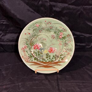May include: A round ceramic plate with a pale green glaze featuring a hand-painted design of pink and red flowers, green foliage, and brown branches. The base of the plate displays a brown decorative border with a cross-hatched pattern.