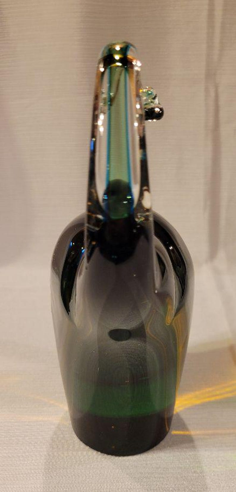 May include: A dark green glass decorative bottle with a long neck and a small handle. The bottle has a rounded base and a clear glass top with blue and green accents. The glass has a glossy finish.