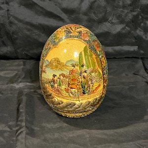 May include: An egg-shaped decorative object with a detailed painted scene of figures in a boat near a mountain. The egg is primarily gold with a textured surface and intricate border detailing. The artwork features various colors.