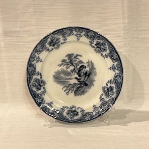 May include: A small, antique white plate with a blue floral border and a central landscape scene. The landscape features a tree, birds, and water. The plate is displayed on a clear stand.