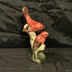 May include: A ceramic figurine of two red birds perched on a branch. The birds have black and brown wing details and are set against a green and cream-colored branch. The base is dark green.