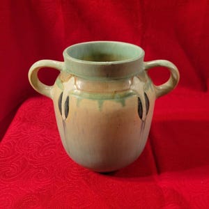 Pu&ograve; includere: Un piccolo vaso in ceramica verde e marrone con due manici. Il vaso ha un design unico con accenti bianchi e neri.