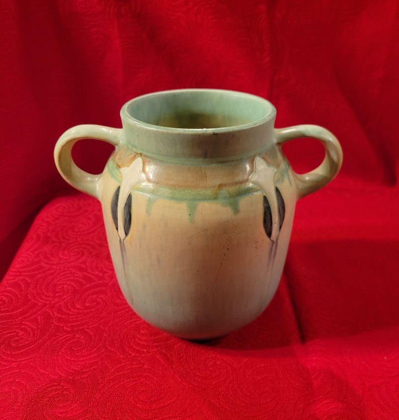 Pu&ograve; includere: Un piccolo vaso in ceramica verde e marrone con due manici. Il vaso ha un design unico con accenti bianchi e neri.