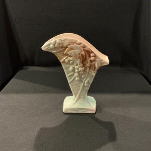 May include: A ceramic vase with a fan-shaped top and a rectangular base. The vase is light pink and green, with a brown grape and leaf design on the front. The vase is approximately 7 inches tall.