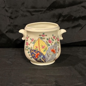 May include: A cream-colored ceramic vase with two handles and a floral design. The vase features colorful leaves in yellow, blue, and gray, with pink flowers and green vines. The rim has a thin blue line.