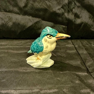 May include: A ceramic figurine of a kingfisher bird. The bird has teal and white coloring with a yellow beak and eye. It is perched on a light green base. The figurine is detailed and appears to be hand-painted.