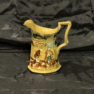May include: A yellow ceramic pitcher with a handle and spout. The pitcher features a painted scene of figures and buildings, with a checkered pattern at the base. The rim and handle are trimmed with gold. A decorative piece for the home.