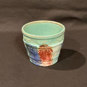 May include: A small, antique ceramic pot with a turquoise interior and a detailed exterior. The pot features a design with blue, red, and white accents. The rim has a gold trim. The pot is likely used for decorative purposes.