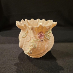 May include: A small, cream-colored ceramic vase with a ruffled rim and a floral design. The vase features a pink rose with green leaves.