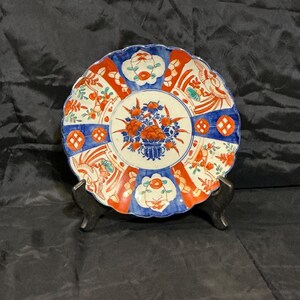 May include: A decorative ceramic plate with a scalloped edge. The plate features a central floral design in orange and blue, surrounded by alternating red and blue segments with floral and geometric patterns. The plate is displayed on a black stand.