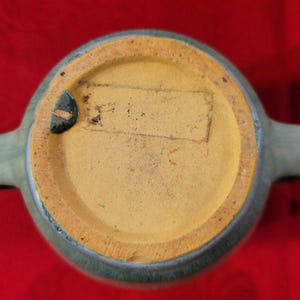 May include: A blue glazed ceramic pitcher with two handles. The bottom of the pitcher has a yellow unglazed base with a label that says "Made in USA".