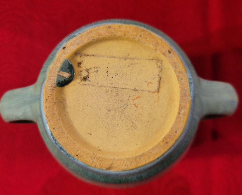 May include: A blue glazed ceramic pitcher with two handles. The bottom of the pitcher has a yellow unglazed base with a label that says "Made in USA".