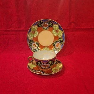 May include: A vintage teacup, saucer, and plate set. The teacup and saucer are placed in front of a larger plate. The set features a floral design with orange, blue, and green accents. The background is a solid red.