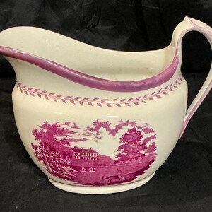 May include: A cream-colored ceramic creamer featuring a magenta transferware landscape scene of a house, trees, and swans. The rim and handle are accented with a magenta border, and a delicate leaf-patterned band encircles the upper body.