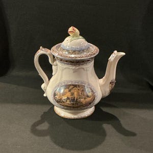 May include: Cream-colored porcelain teapot with a detailed handle and spout. The lid is topped with a flower-shaped knob. The body features a painted oval scene with figures in warm tones, and is trimmed with blue and brown decorative bands. A classic tea set item.