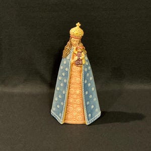 May include: A ceramic figurine of a woman wearing a gold crown and a blue cloak adorned with white floral patterns. She holds a smaller figure, with a peach-colored dress and a gold patterned dress. The figurine has a peach-colored dress with a gold patterned design.