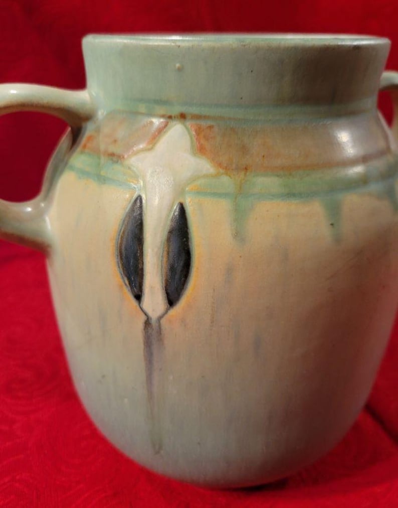 Pu&ograve; includere: Un vaso in ceramica verde smaltato con due manici e un design floreale stilizzato in bianco, marrone e nero.