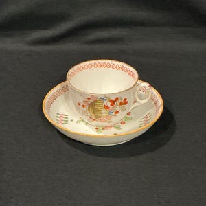 May include: A white teacup and saucer set with a floral design. The cup and saucer have a gold trim and a red and gold border. The floral design includes red, pink, and blue flowers.