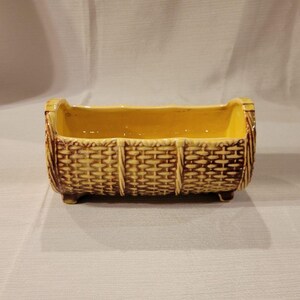 May include: A yellow ceramic planter shaped like a woven basket with brown accents. The planter has four small feet and is designed to hold a small plant.