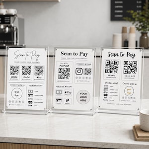 Puede incluir: Tres soportes verticales de acrílico transparente que muestran tarjetas informativas blancas con texto y gráficos en negro. Cada tarjeta presenta encabezados Scan to Pay, múltiples códigos QR para servicios de pago e iconos de redes sociales.