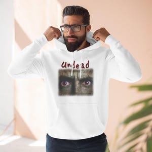 May include: White hooded sweatshirt with the word "Undead" in maroon script above a graphic of red eyes. The graphic is a close-up of a face with a dark, textured background. The sweatshirt has a drawstring hood and a front pocket.