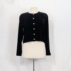 May include: A black velvet cardigan with long sleeves and a round neckline. The cardigan is fastened with six iridescent buttons. The garment is displayed on a mannequin against a white backdrop.