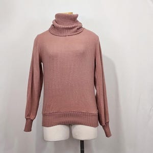 Könnte beinhalten: Ein altrosa Rollkragenpullover mit langen Ärmeln. Der Pullover hat einen gerippten Kragen, Bündchen und Saum. Die Ärmel sind an den Schultern leicht aufgebauscht. Der Pullover wird auf einer Schaufensterpuppe präsentiert.
