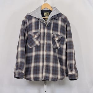 Puede incluir: Una camisa de franela a cuadros gris, azul y blanco con capucha gris. La camisa tiene dos bolsillos en el pecho y una parte delantera abotonada. La camisa está colgada de una percha de madera.