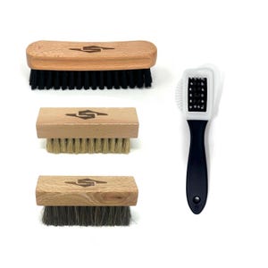Könnte beinhalten: Eine Sammlung von Schuhreinigungsbürsten. Drei Holzbürsten mit unterschiedlich farbigen Borsten: schwarz, beige und grau. Jede Bürste hat einen Holzgriff mit einem Logo. Eine schwarz-weiße Schuhbürste mit schwarzem Griff ist ebenfalls enthalten.