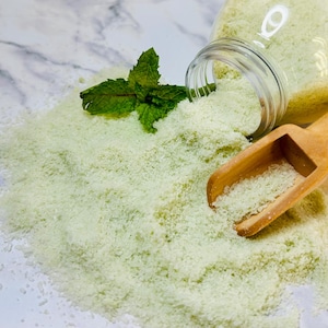 May include: A clear glass jar tipped over, spilling pale green granules onto a marble surface. A small wooden scoop and fresh mint leaves are next to the granules. The granules appear to be a bath product.