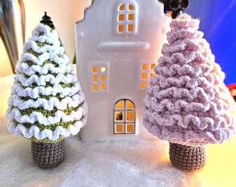May include: Two crocheted Christmas trees, one green and white and one pink, stand in front of a white ceramic house with windows.