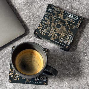 May include: Black ceramic mug with coffee on a black circuit board coaster. The coaster has gold-colored circuit designs and the text "Crafted for the curious." Another circuit board coaster is next to the mug. A laptop is in the background.