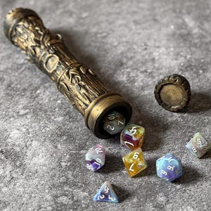 Könnte beinhalten: Ein bronzefarbener Würfelbehälter mit einem verzierten, strukturierten Design, der geöffnet ist, um mehrere farbenfrohe polyedrische Würfel zu enthüllen. Die Würfel sind in verschiedenen Farben gehalten, darunter Blau, Lila und Gelb. Der Behälter ist zylindrisch und hat eine abnehmbare Kappe.