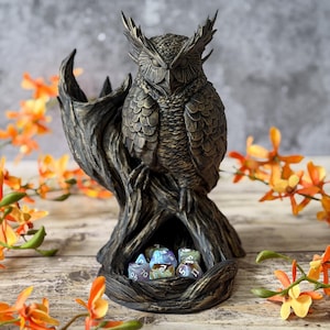 May include: A bronze owl ornament, intricately detailed with a tree trunk base, holding several multi-sided dice. The owl has closed eyes and detailed feather textures. The background features orange flowers.