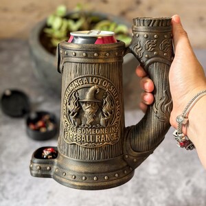 Op de afbeelding: Een donkerbronskleurige drankblikhouder in de vorm van een pul met een houtnerftextuur. Het heeft een reliëf van een tovenaar die vlammen vasthoudt en de tekst "TALKING A LOT OF SHIT FOR SOMEONE IN FIREBALL RANGE." Inclusief een klein zijvakje.