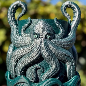 Könnte beinhalten: Ein teal-farbener Pflanztopf mit einem detaillierten Oktopus-Design. Der Oktopus hat silberne Akzente, grüne Augen und Tentakel, die sich um den Pflanztopf wickeln. Die Griffe werden von den Oktopus-Tentakeln gebildet. Geeignet für den Innen- oder Außenbereich.