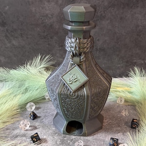 May include: A metallic green dice tower with intricate embossed patterns and a geometric top. A small, square tag with a relief design hangs from a rope detail. Several dice are scattered around the base of the tower.