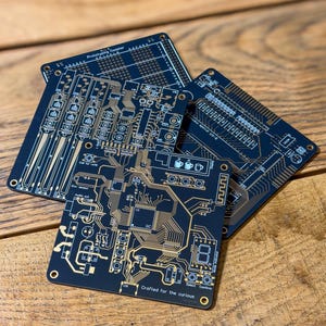 Op de afbeelding: Vier zwarte onderzetters met printplaatontwerp en goudkleurige elektrische paden en componenten. Op een staat "Prototyping Coaster". De onderste onderzetter heeft de tekst "Crafted for the curious".