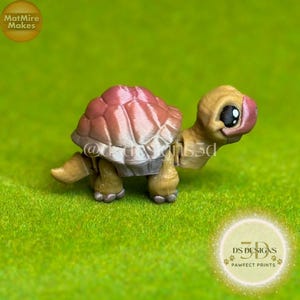 May include: A 3D-printed turtle figurine with a tan head and limbs, a pink and brown shell, and a white underbelly. The turtle is set against a green background. The figurine has large black eyes and is a decorative item.